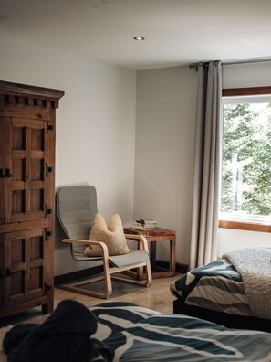 Una habitación con un armario de madera, un sillón gris con cojín, una pequeña mesa de madera con libros y una cama con manta estampada.
