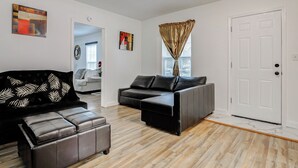 Een moderne woonkamer met een hoekbank, een tweezitterbank en een bijpassend voetenbankje. Aan de rechterkant is een deur en er is een raam met jaloezieën.