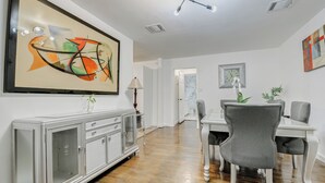 Een eethoek met een witte tafel, grijze stoelen, een kast met glazen deuren en een groot abstract schilderij aan de muur.