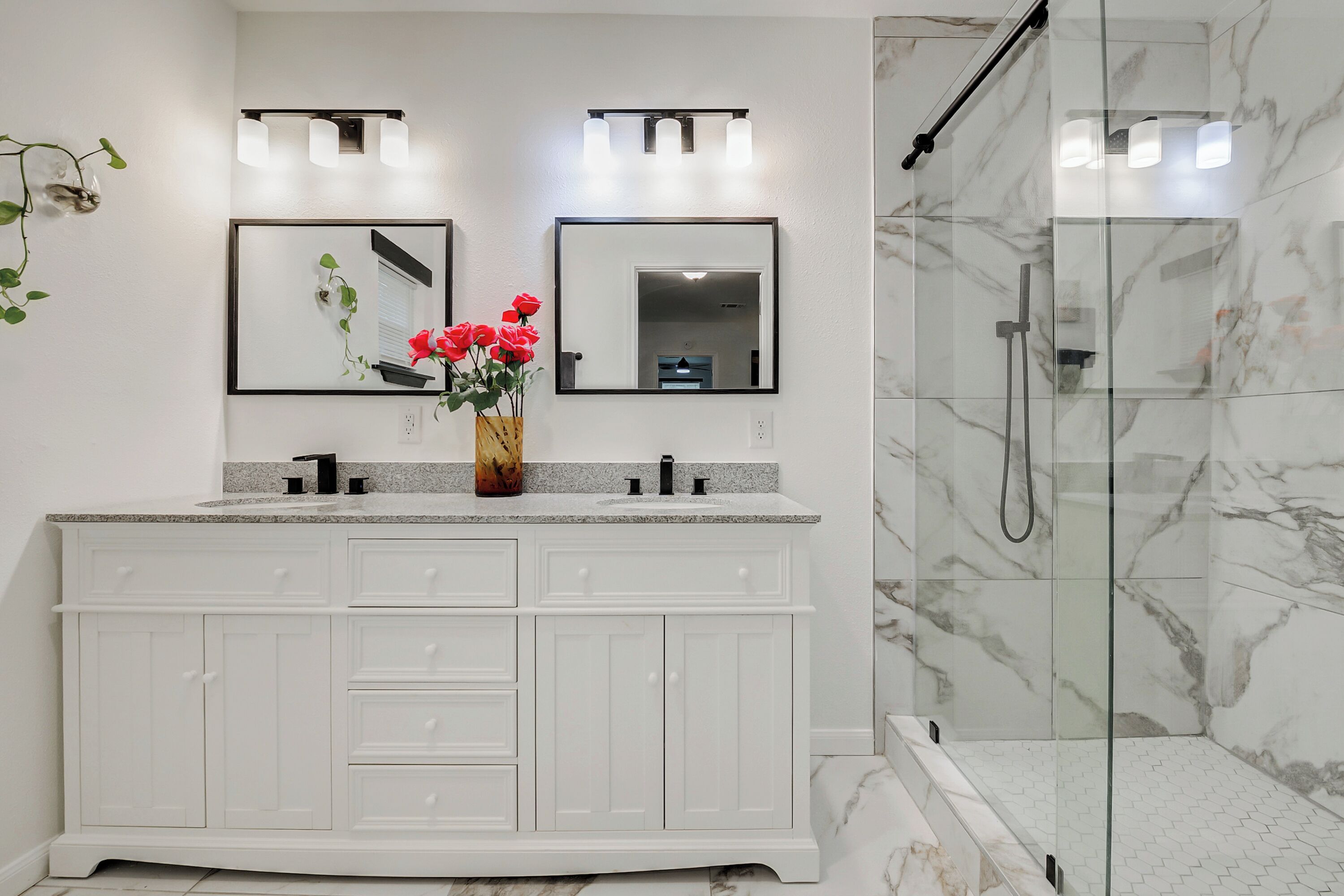 Une salle de bain avec un meuble-lavabo blanc, deux miroirs, un vase avec des fleurs et une cabine de douche en verre.