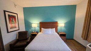 Een hotelkamer met een bed, twee bedlampen, een stoel, een inlijsting foto aan de muur, en een raam met gordijnen.