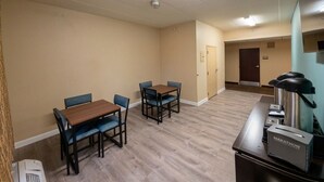 Een hotelkamer met een houten tafel en stoelen, een kitchenette met een koffiezetapparaat, en een deur die leidt naar een andere kamer.