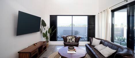 A modern living room with a large flat-screen TV, leather sofas, a coffee table, and a large window with blinds.