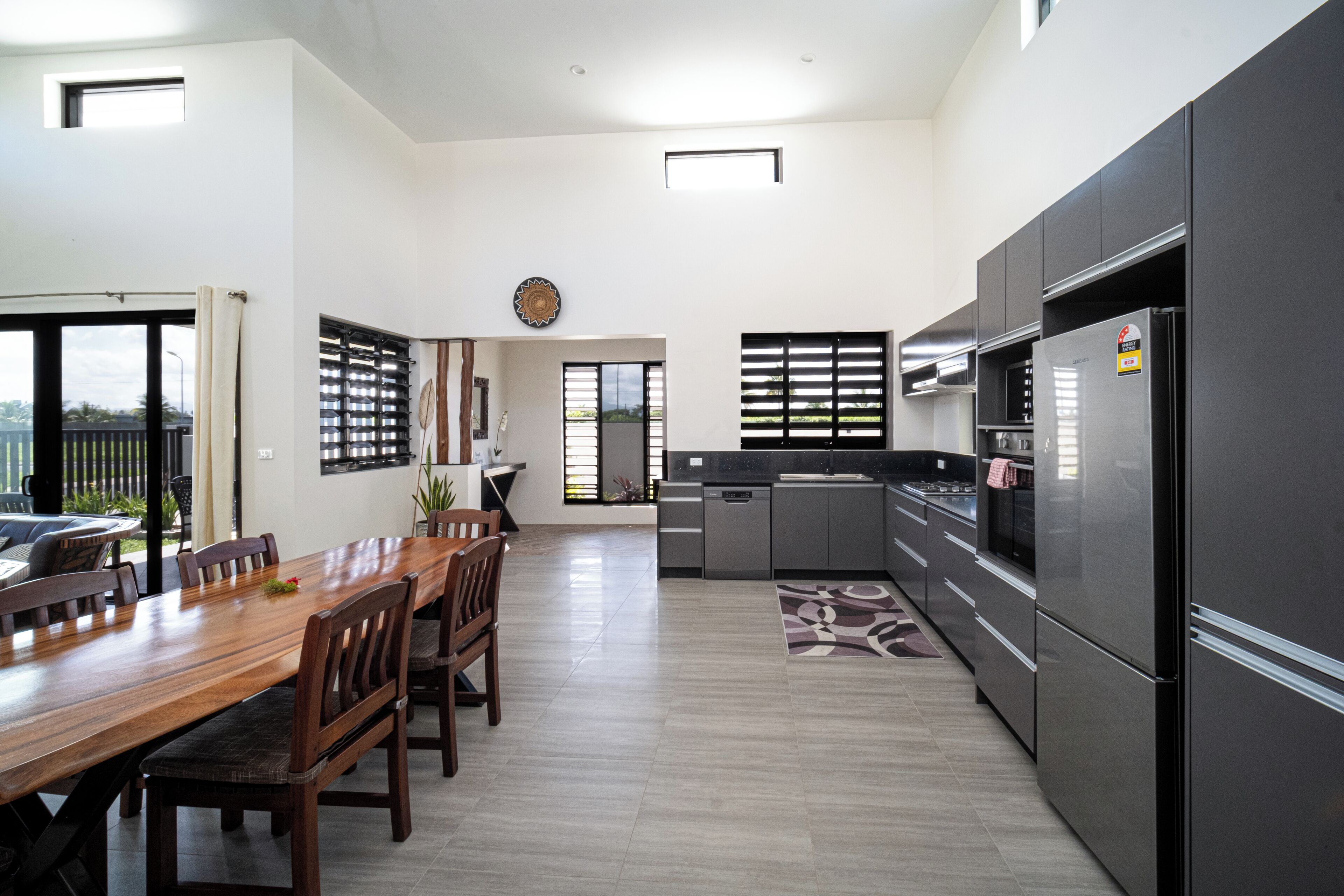 A modern kitchen with a dining area, featuring a large wooden table, dark cabinets, and a view through the windows.