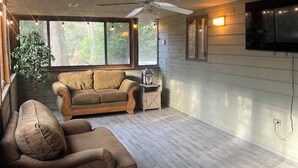Un salón amplio con ventilador de techo, un ventanal grande, televisor montado en la pared y suelo de madera.