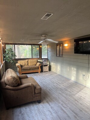 A spacious living room with a ceiling fan, a large window, a television mounted on the wall, and a wooden floor. - Family-friendly, Spacious, One-Level 4-Bedroom Home in Tranquil Southern Shores (Kitty Hawk)