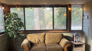 Una habitación con piso de madera, un sofá marrón, una planta en maceta, una ventana con vista a árboles y un pequeño mueble de madera con una radio.