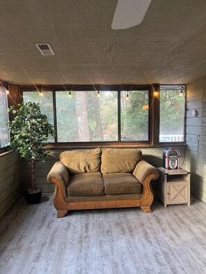 A room with a wooden floor, a brown sofa, a potted plant, a window with a view of trees, and a small wooden cabinet with a radio. - Family-friendly, Spacious, One-Level 4-Bedroom Home in Tranquil Southern Shores (Kitty Hawk)