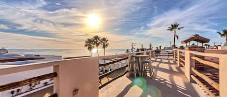 A beachside terrace with a view of the ocean, palm trees, and a wooden fence.