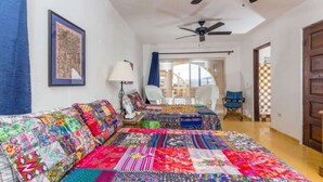 Une chambre avec deux lits recouverts de couvertures aux couleurs vives, un ventilateur de plafond, un tableau encadré au mur et une vue sur un balcon par la fenêtre.