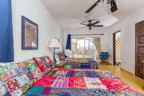 Ein Zimmer mit zwei Betten, die mit bunten Quilts bezogen sind, einem Deckenventilator, einem gerahmten Bild an der Wand und Blick auf einen Balkon durch ein Fenster.