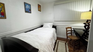 Un lit simple avec une couette blanche, une chaise en bois, un bureau avec une lampe et deux tableaux accrochés au mur.