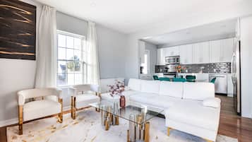 Ein modernes Wohnzimmer mit einem Glastisch, weißen Sofas und einer Küche im Hintergrund.