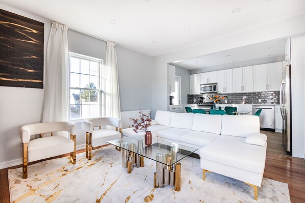 A modern living room with a glass coffee table, white sofas, and a kitchen in the background.