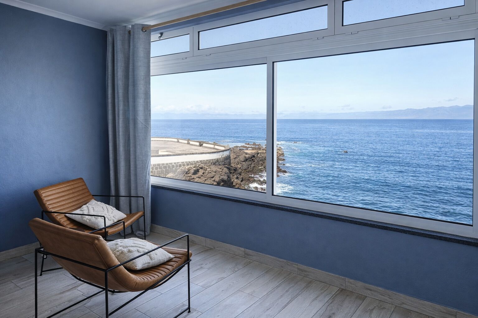 A room with a large window, a chair, and a view of the sea and rocky coastline.