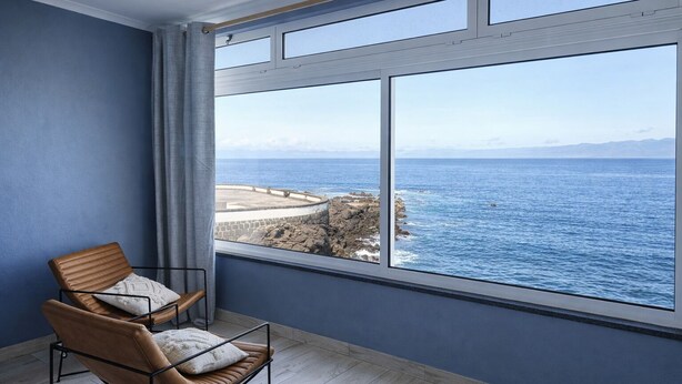 Une pièce dotée d’une grande fenêtre, d’un fauteuil et offrant une vue sur la mer et la côte rocheuse.