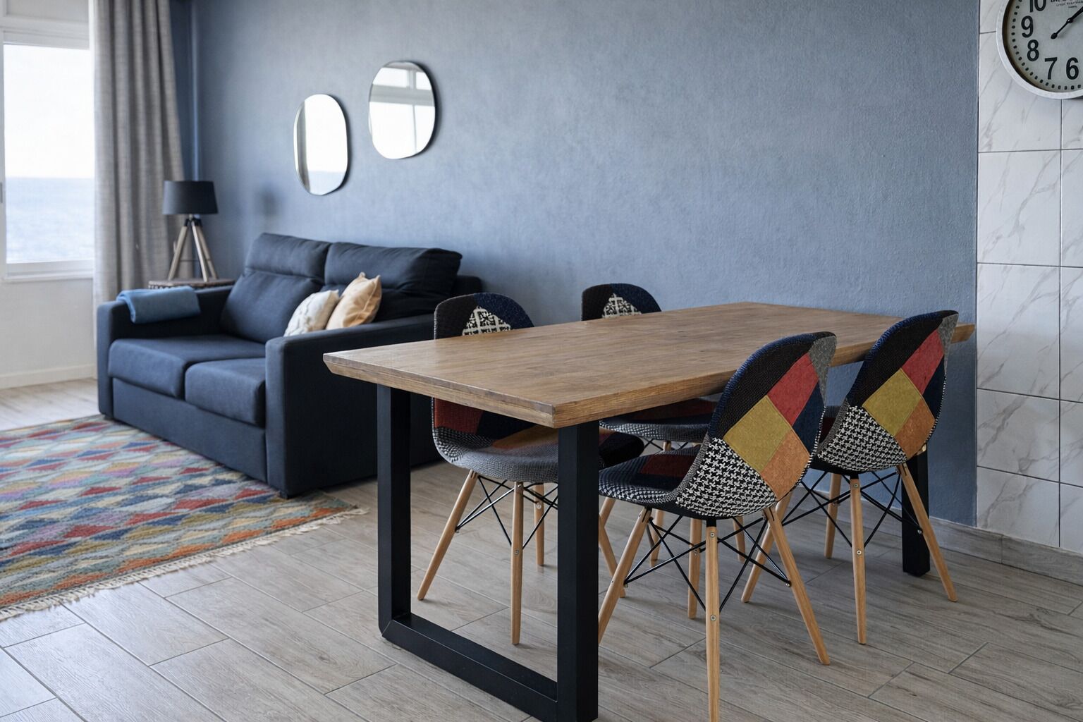 A modern living room with a dark sofa, a wooden dining table, and patterned chairs.