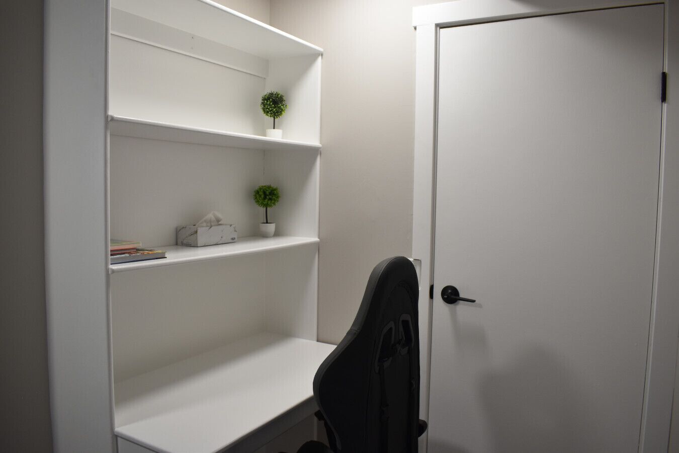 Una puerta blanca con picaporte negro, un estante blanco con una planta pequeña y una silla de oficina negra.