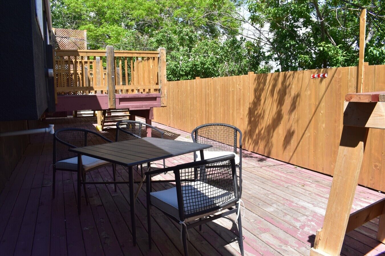 En udendørs terrasse med et bord og stole, et træhegn og en træbygning i baggrunden.
