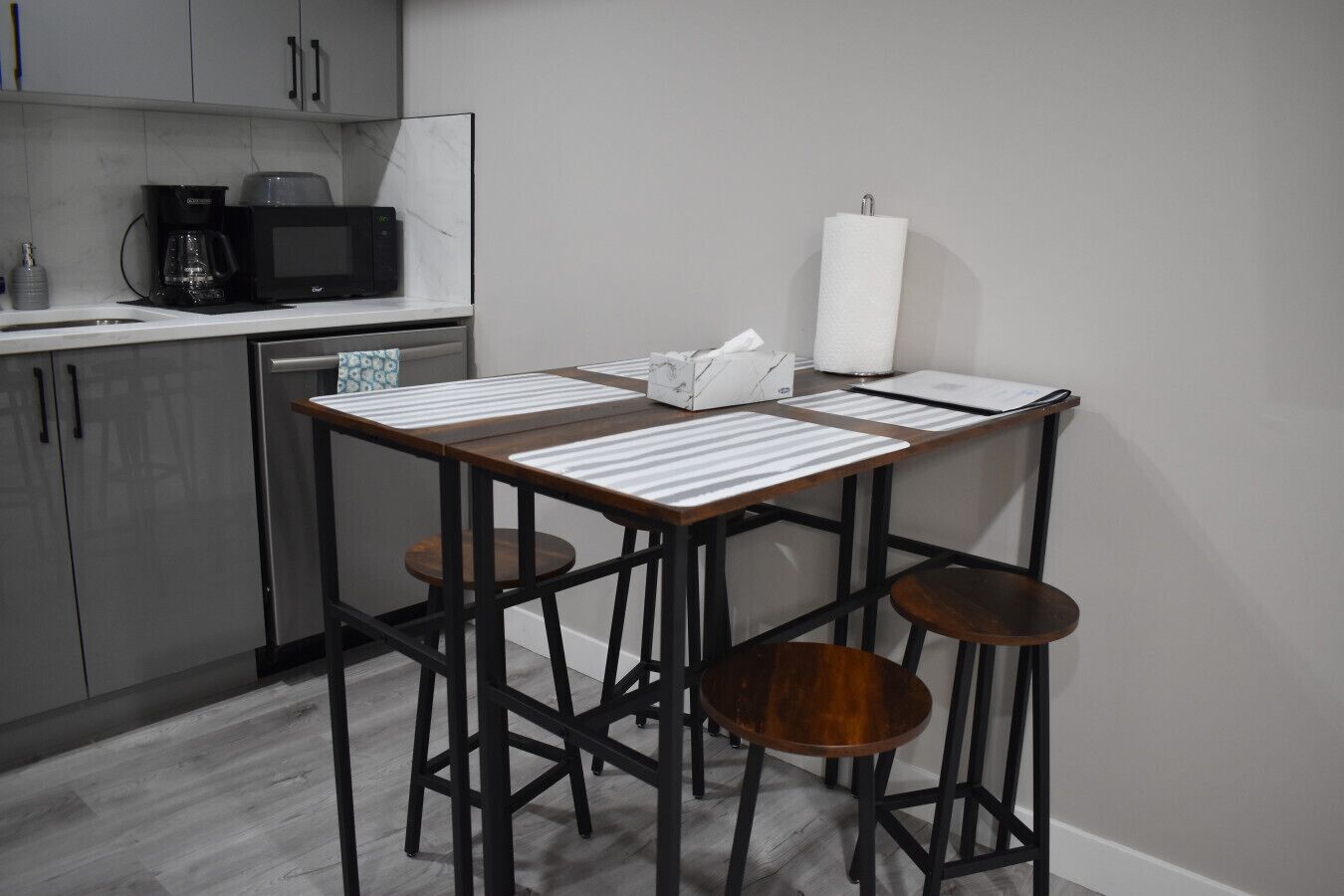 Eine kompakte Küche mit Mikrowelle, Kaffeemaschine und Küchenpapierrolle. Zwei Hocker und ein kleiner Tisch mit gestreiften Tischsets.