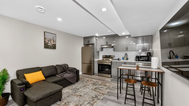 Una cocina moderna con electrodomésticos de acero inoxidable, una zona de comedor con taburetes de bar y un sofá gris con un cojín amarillo.