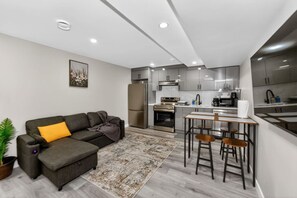 Una cocina moderna con electrodomésticos de acero inoxidable, una zona de comedor con taburetes de bar y un sofá gris con un cojín amarillo.