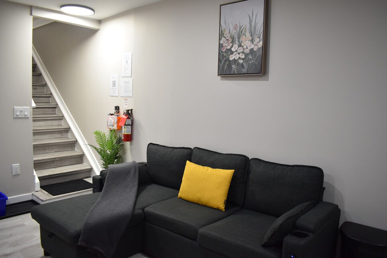 Ein Wohnzimmer mit grauem Sofa, einem gelben Kissen, einem Feuerlöscher und einer Treppe.