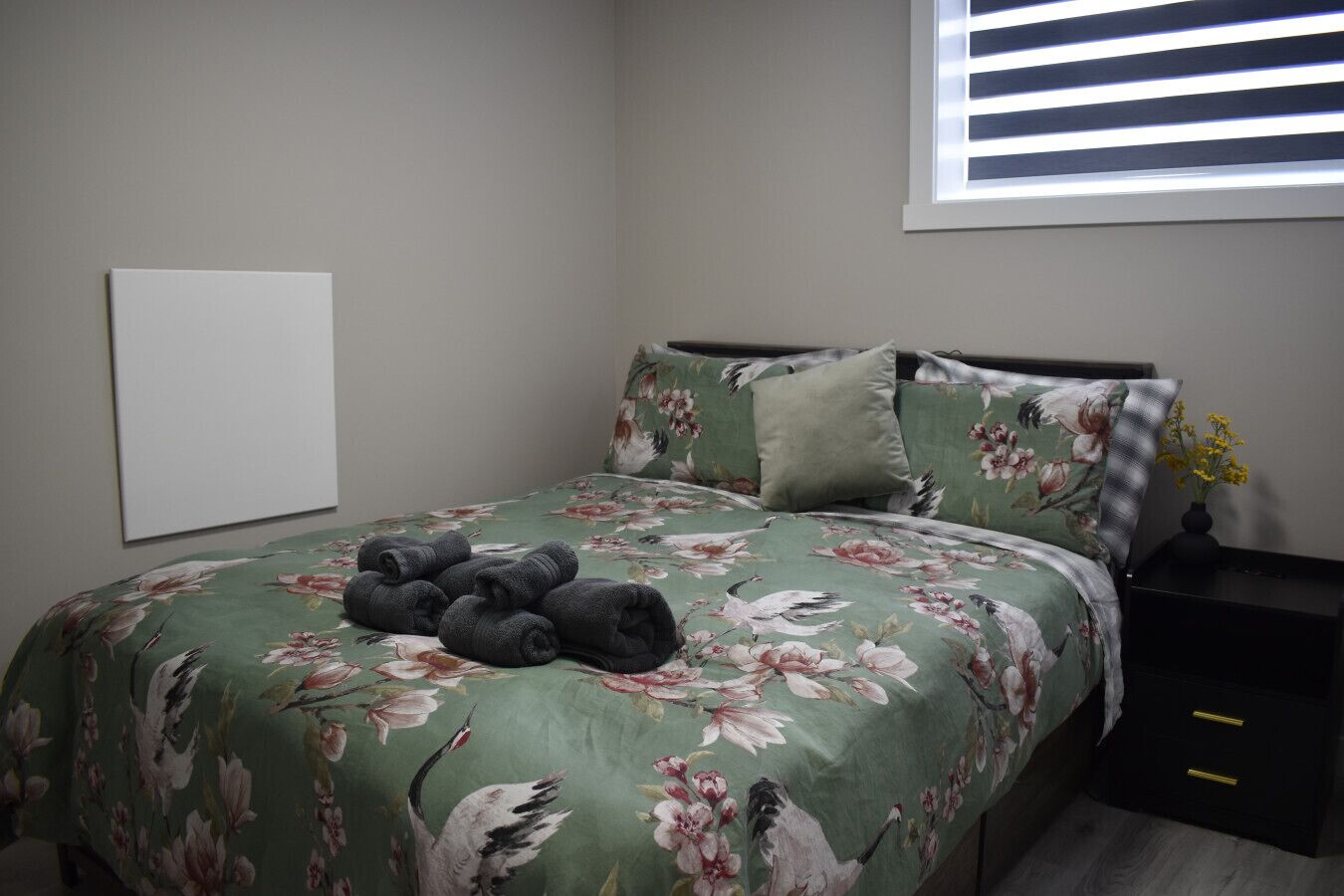 A bedroom with a floral bedspread, a neatly made bed with pillows, a bedside table with a vase, and a window with blinds.