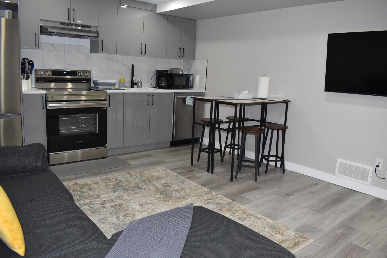 A modern kitchen with stainless steel appliances, a dining area with a table and bar stools, a gray sofa, and a flat-screen TV mounted on the wall.