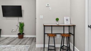 Una habitación con una televisión de pantalla plana montada en la pared, una mesita con dos taburetes, una planta en maceta y una puerta.