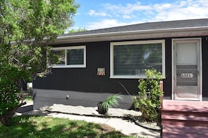 A single-story house with a dark exterior, a front door with a number plaque, and a small garden area with a tree and plants.