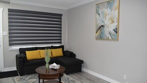Een woonkamer met een zwarte bank, een ronde houten salontafel, een kamerbreed tapijt met een patroon, een groot abstract schilderij en een raam met horizontale jaloezieën.