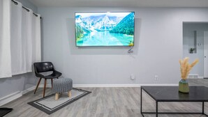 Una sala moderna con televisor de pantalla plana, una silla negra, un reposapiés gris, una mesa de centro negra y un jarrón con plantas secas.