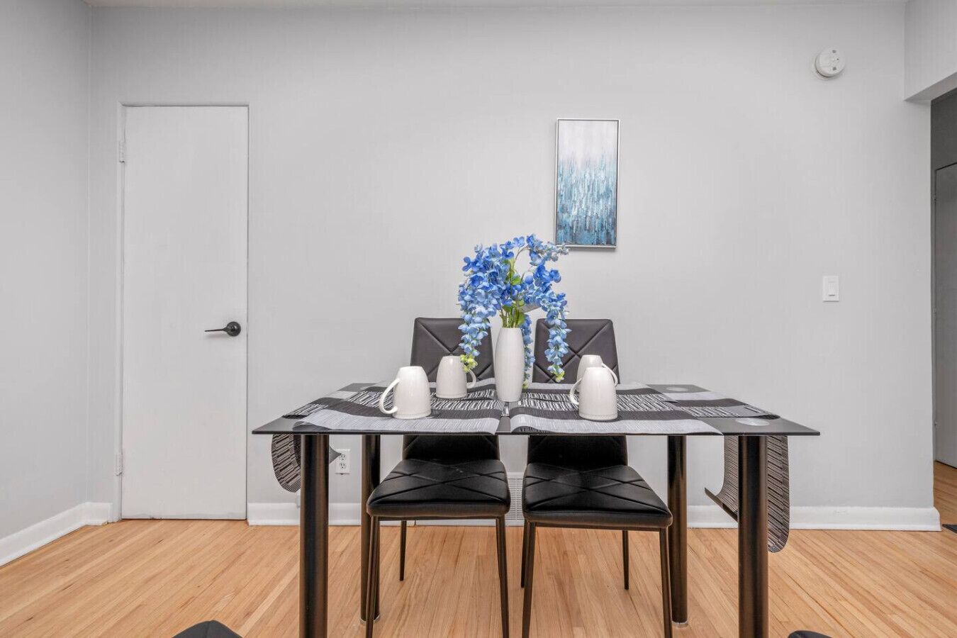 Een eetgedeelte met een tafel gedekt voor twee personen, twee stoelen, een vaas met blauwe bloemen en een schilderij aan de muur.