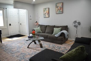Un salon avec un canapé, une table basse en verre, un tapis, un ventilateur et une porte.