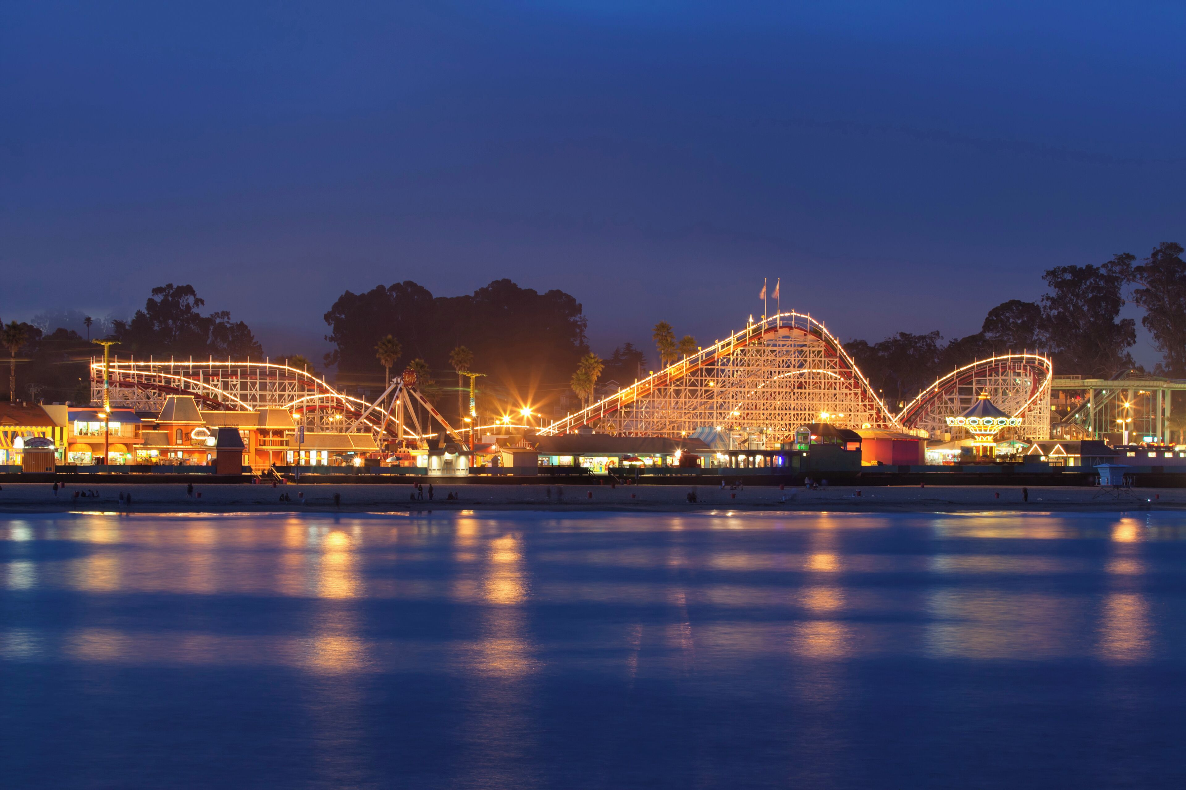 A nighttime view of a water park with illuminated roller coasters and a calm water surface reflecting the lights.