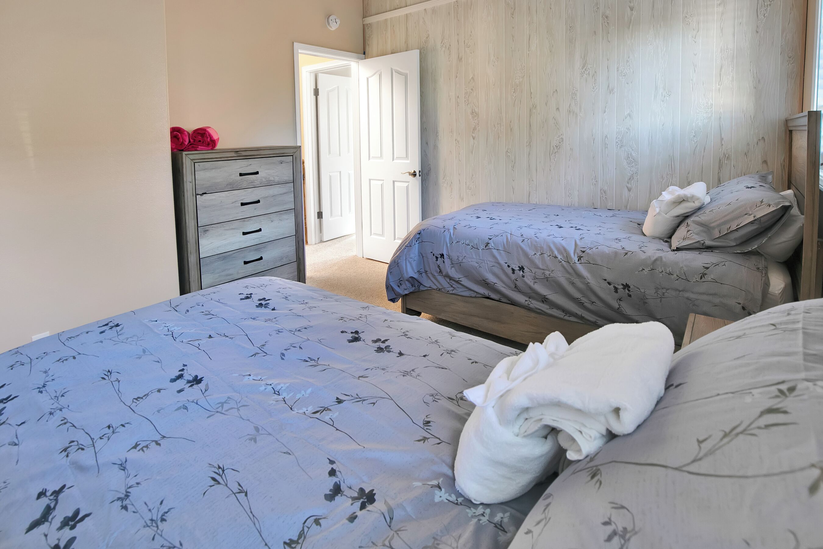 Une chambre avec deux lits, une commode en bois et une porte donnant sur une autre pièce.