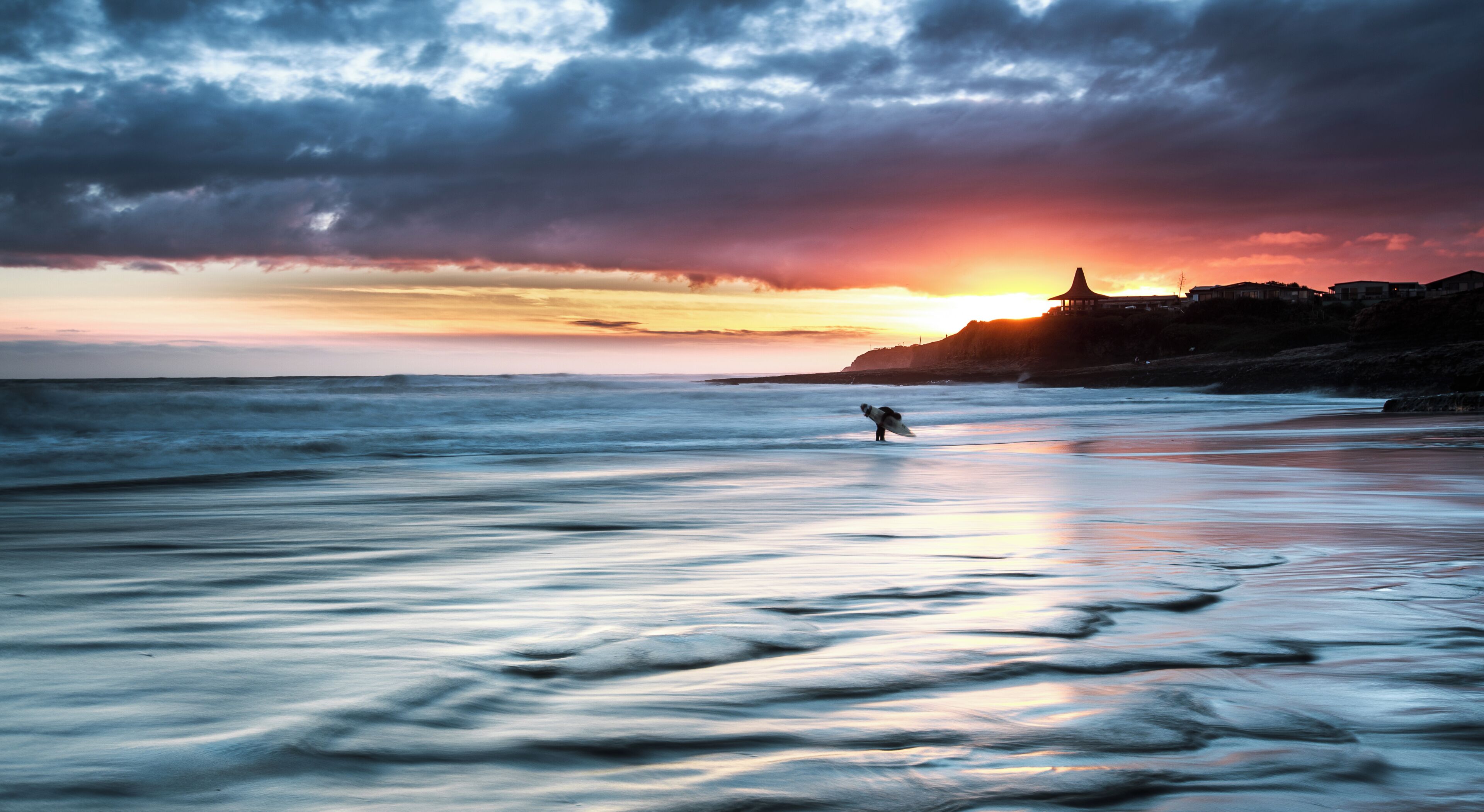 A beach with a sunset, calm water, and a person surfing.