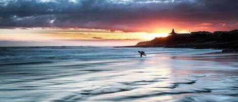 A beach with a sunset, calm water, and a person surfing.
