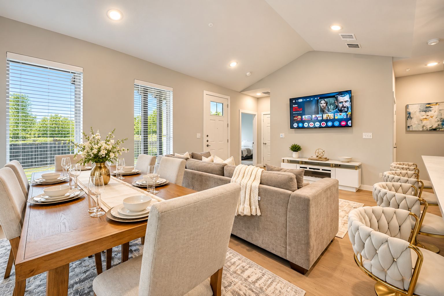 A modern living room with a dining area, a flat-screen TV mounted on the wall, and a large window with blinds.