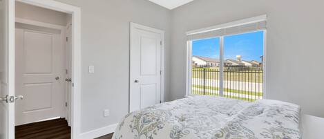 Une chambre à coucher avec un grand lit, deux portes blanches et une fenêtre donnant sur une clôture et des immeubles.