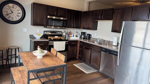 A modern kitchen with dark wood cabinets, stainless steel appliances, a wooden dining table, and a wall clock.