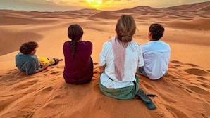 Four people sitting on sand dunes facing a sunset.