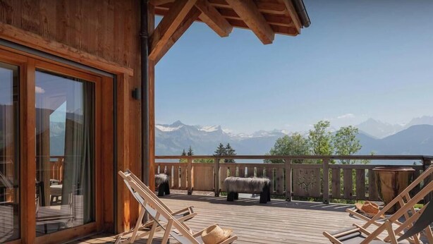 Une terrasse en bois avec des chaises longues, une porte vitrée et une vue sur des montagnes enneigées.