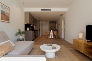 A modern living room with a sofa, a coffee table, a TV stand, and a dining area in the background. - Premium Apartment, Poolview, King + Queen (Sitges)