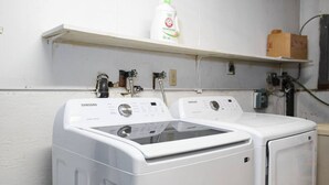 A laundry area with two Samsung washing machines, a shelf with a bottle of laundry detergent, and a cardboard box. - Cozy Bear Den Rustic Cabin Central Located (Fairbanks)