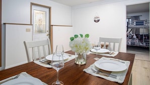 Un coin repas avec une table en bois dressée pour quatre personnes, des chaises blanches, un vase avec des fleurs blanches et un tableau encadré au mur.