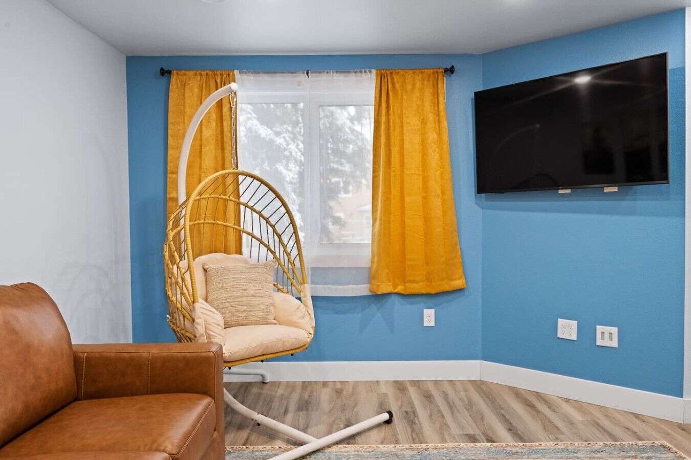 A room with a blue wall, a television mounted on the wall, a hanging chair, and a brown leather sofa.
