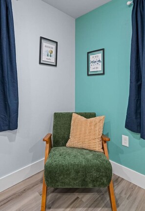 Una habitación de esquina con paredes verde azuladas, un sillón tapizado en verde con un cojín color beige, y dos cuadros enmarcados en las paredes.