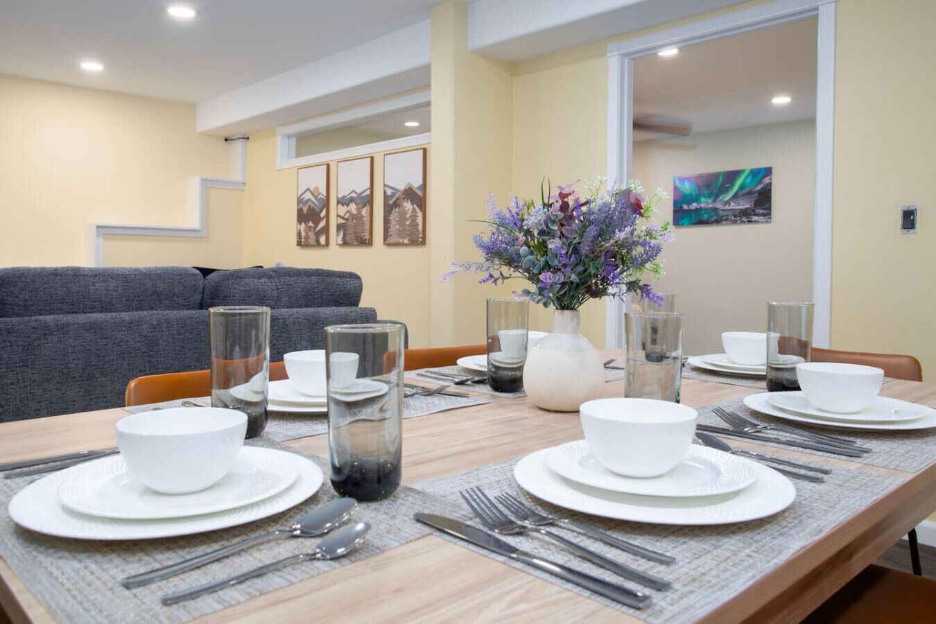 A dining area with a wooden table set for four, white plates, cutlery, and clear glassware. A vase with purple flowers is on the table.